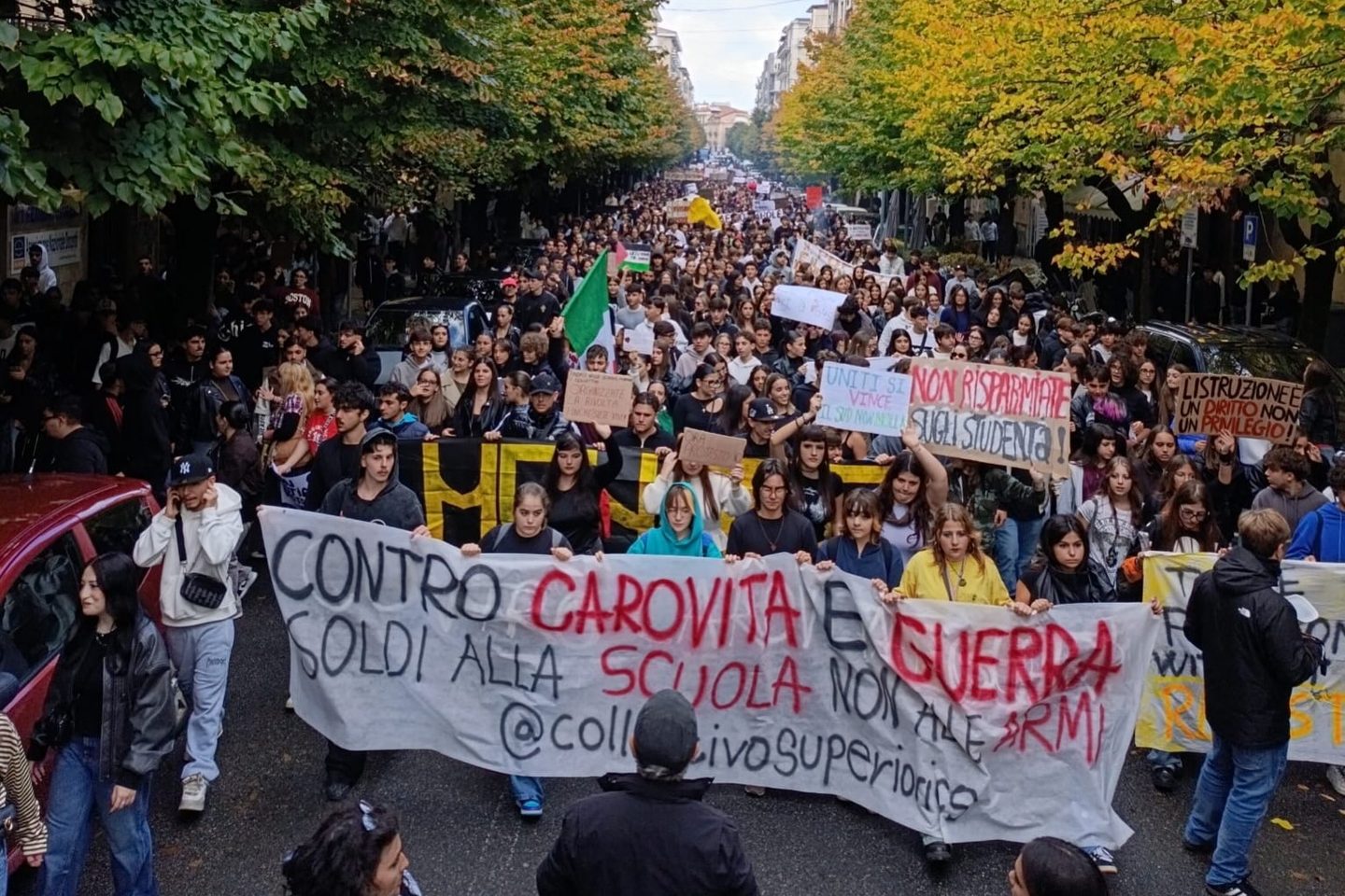 Il corteo degli studenti a Cosenza, 22 ottobre 2026: in testa lo striscione «Contro carovita e guerra – Soldi alla scuola non alle armi», dietro centinaia di ragazzi con cartelli tra due file di alberi.